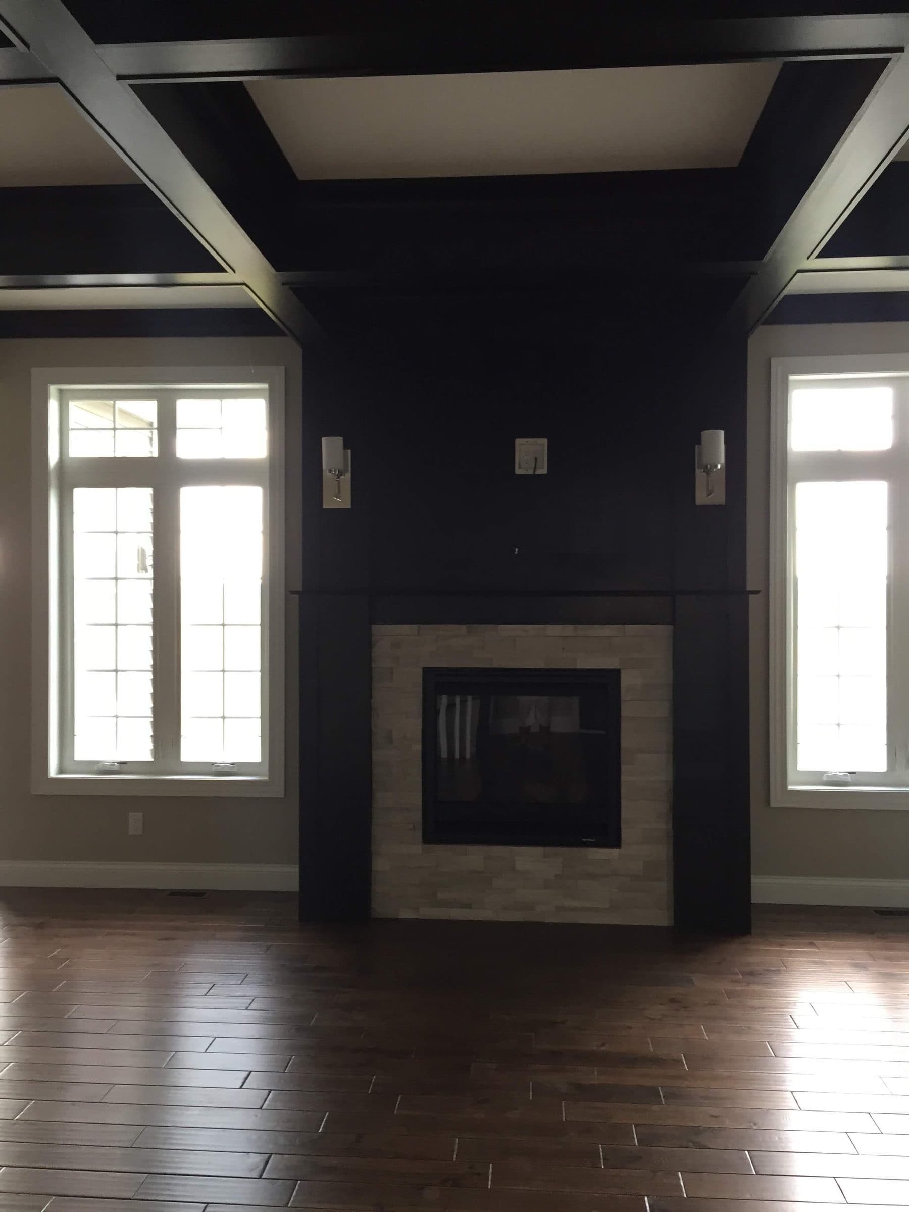 Dark-painted coffered ceiling carried down onto the fireplace wall, framed by white windows and dark hardwood floors.