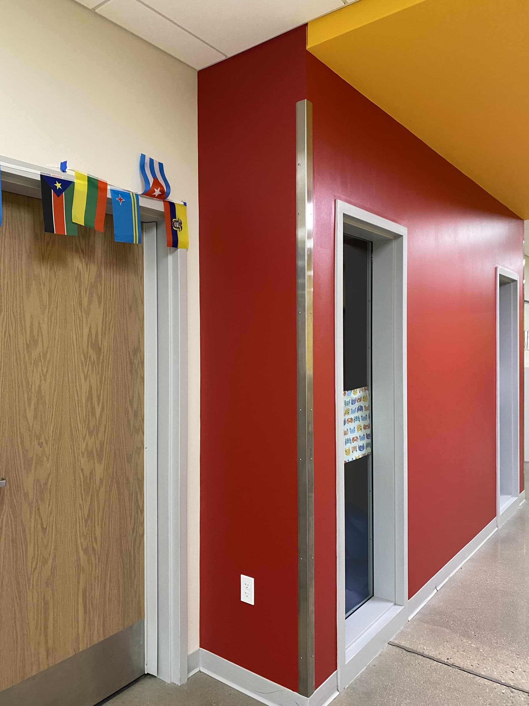 School corridor with red and yellow accent walls meeting at a corner above a wood door and a row of coat hooks.