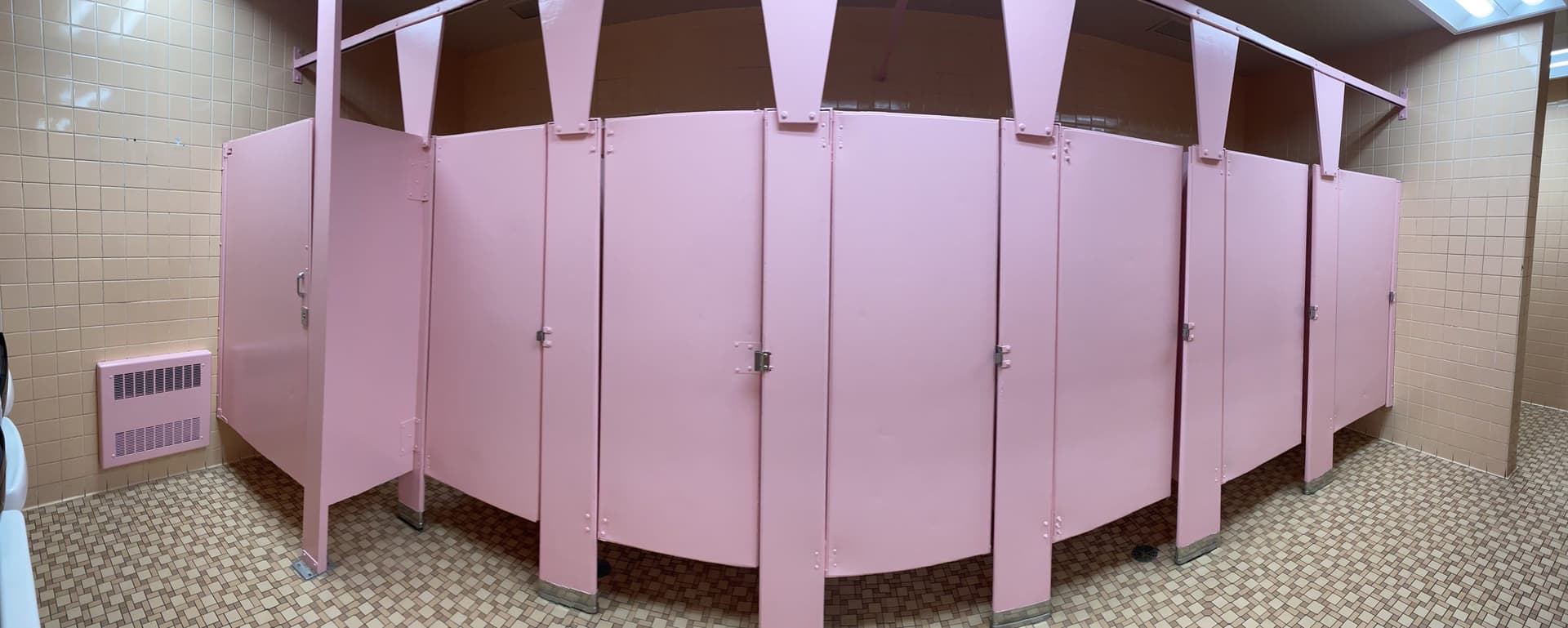 School restroom partitions recoated in pink against white tile, photographed as a wide panorama.