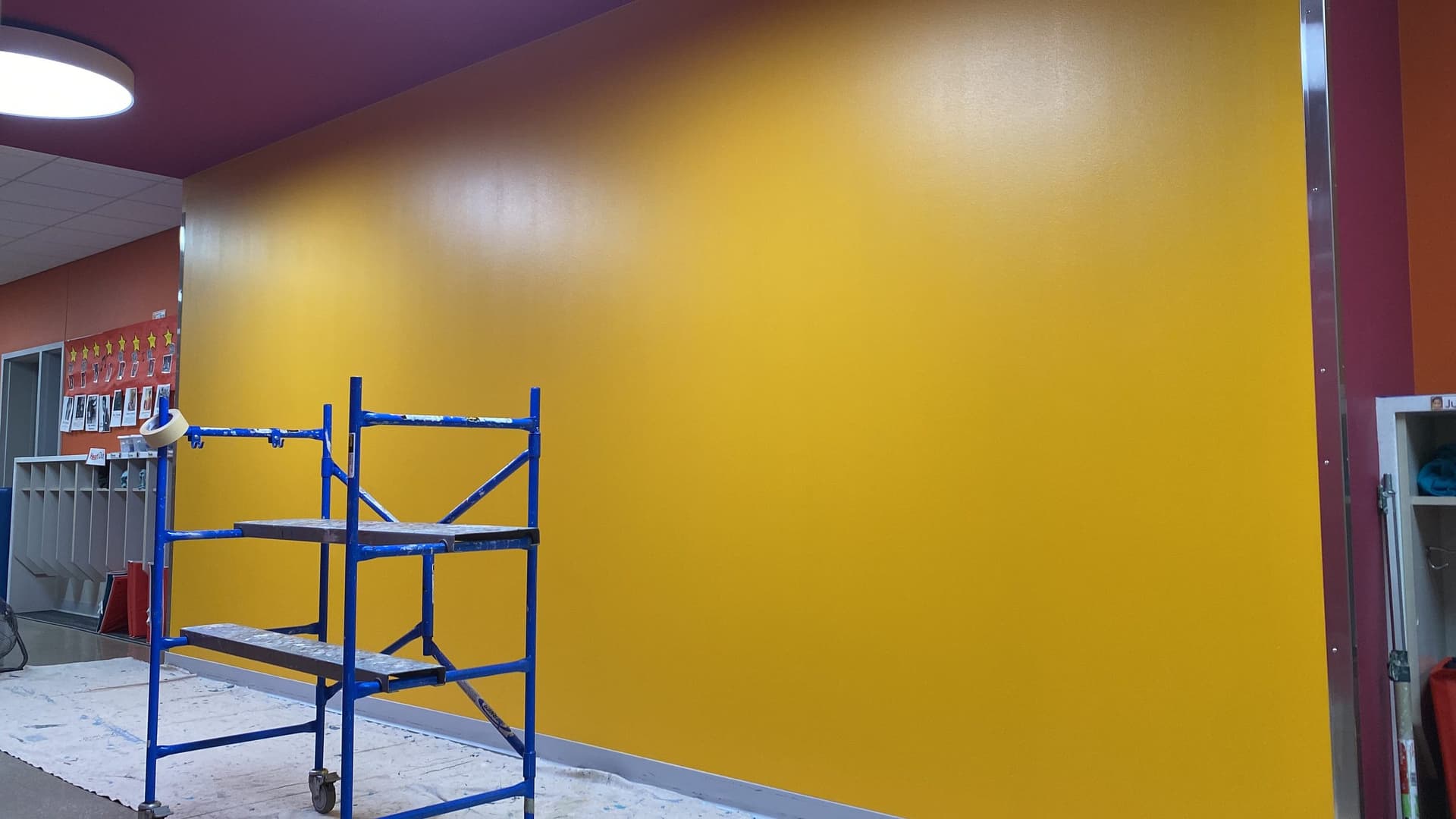 Large yellow wall rolled out from a mobile scaffold tower in a school commons, with a red wall beyond.