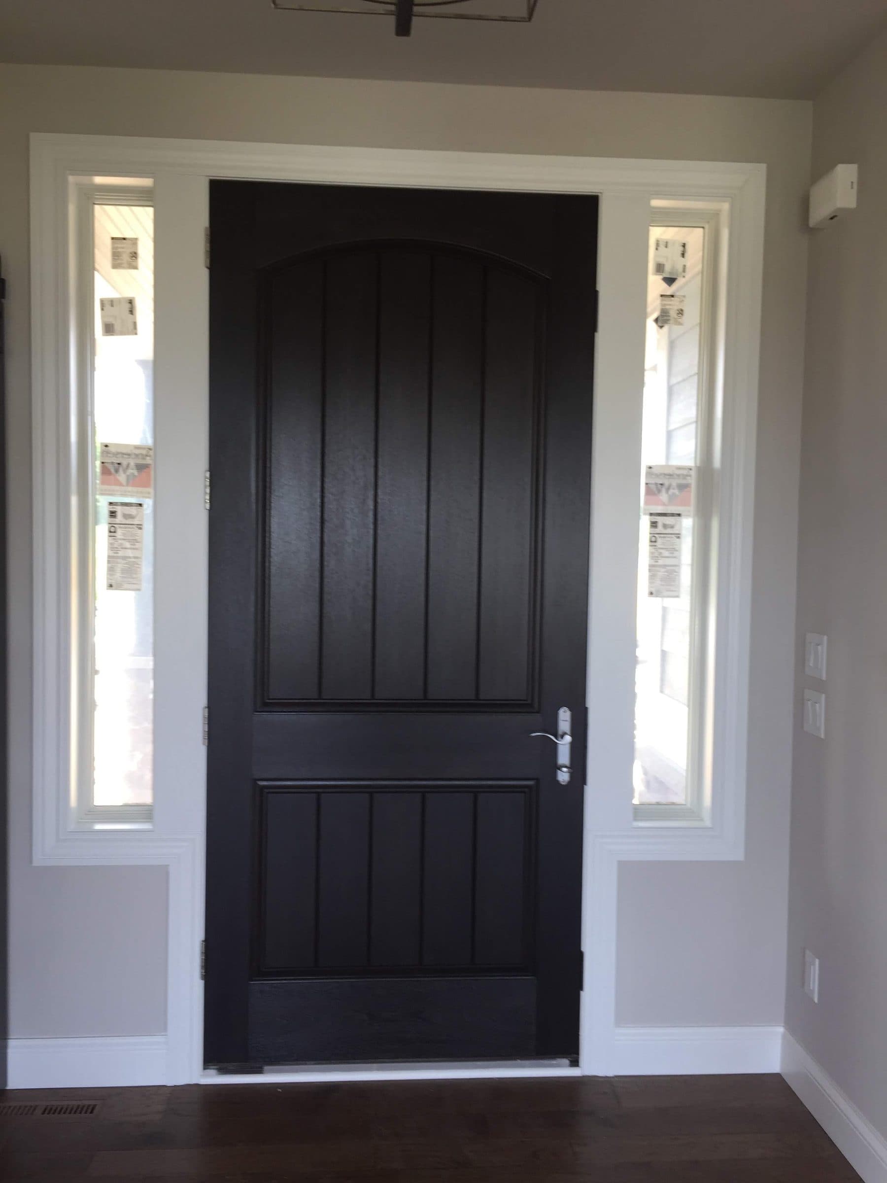 Black-stained fiberglass entry door with an arched plank panel, flanked by two frosted sidelights, seen from inside against white trim and dark hardwood floors.