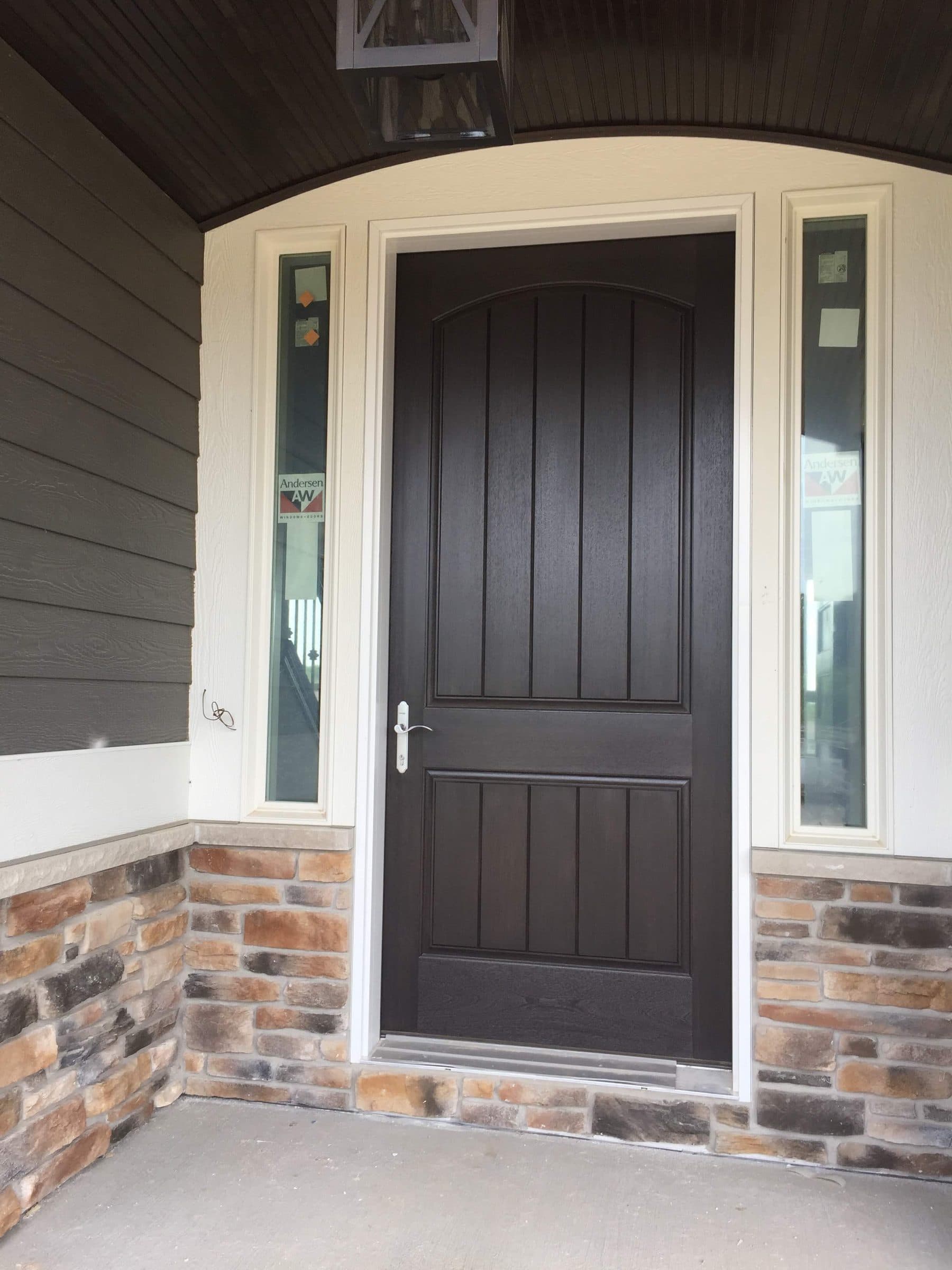 The same black-stained entry door from the porch, set in gray lap siding above a stone veneer base under a curved bead-board porch ceiling.