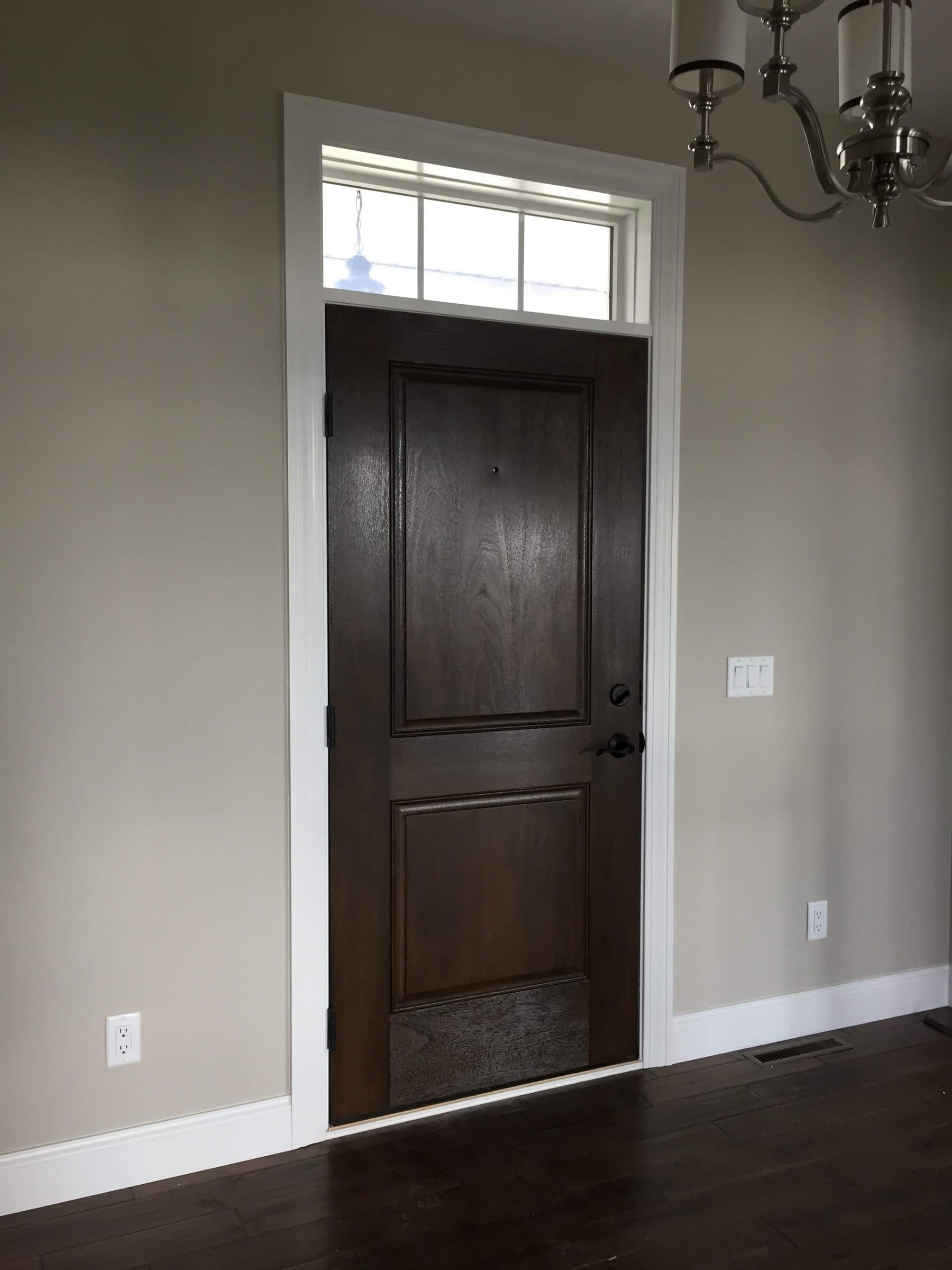 Dark-stained interior door with a clear transom window above it, set in white casing against warm gray walls.