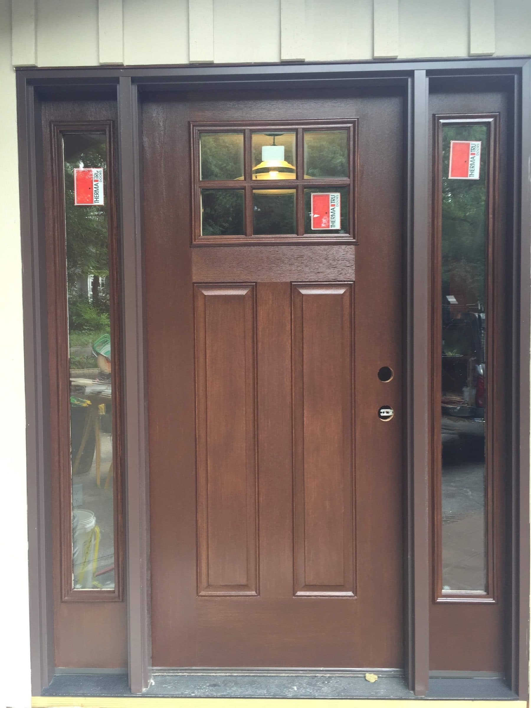 Brown-stained craftsman entry door with a four-light top rail and two sidelights, photographed on a new build with the manufacturer tags still on the glass.