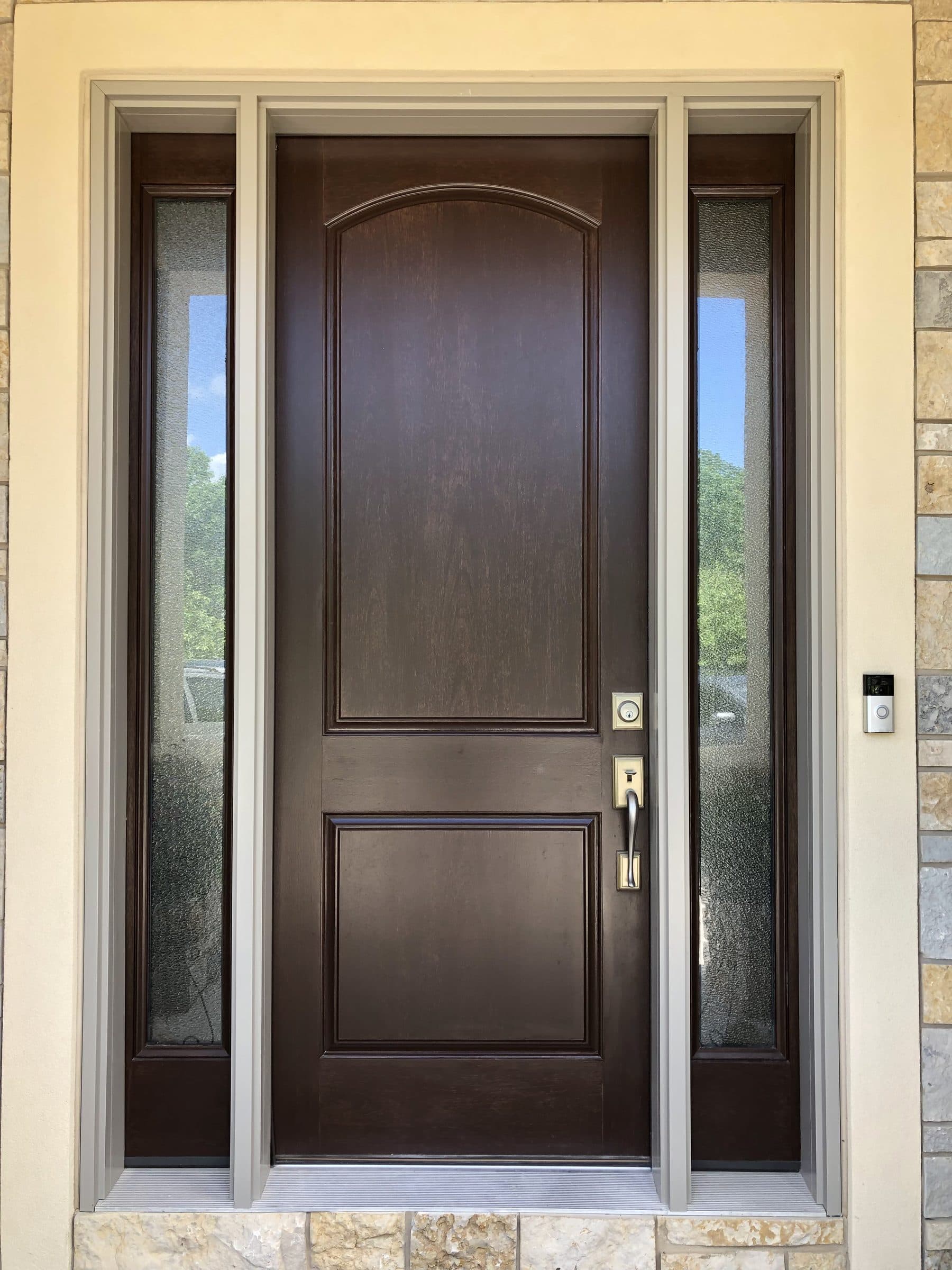 Warm brown-stained entry door with a raised arch panel and two divided-light sidelights, set in cream stucco above a stone base.