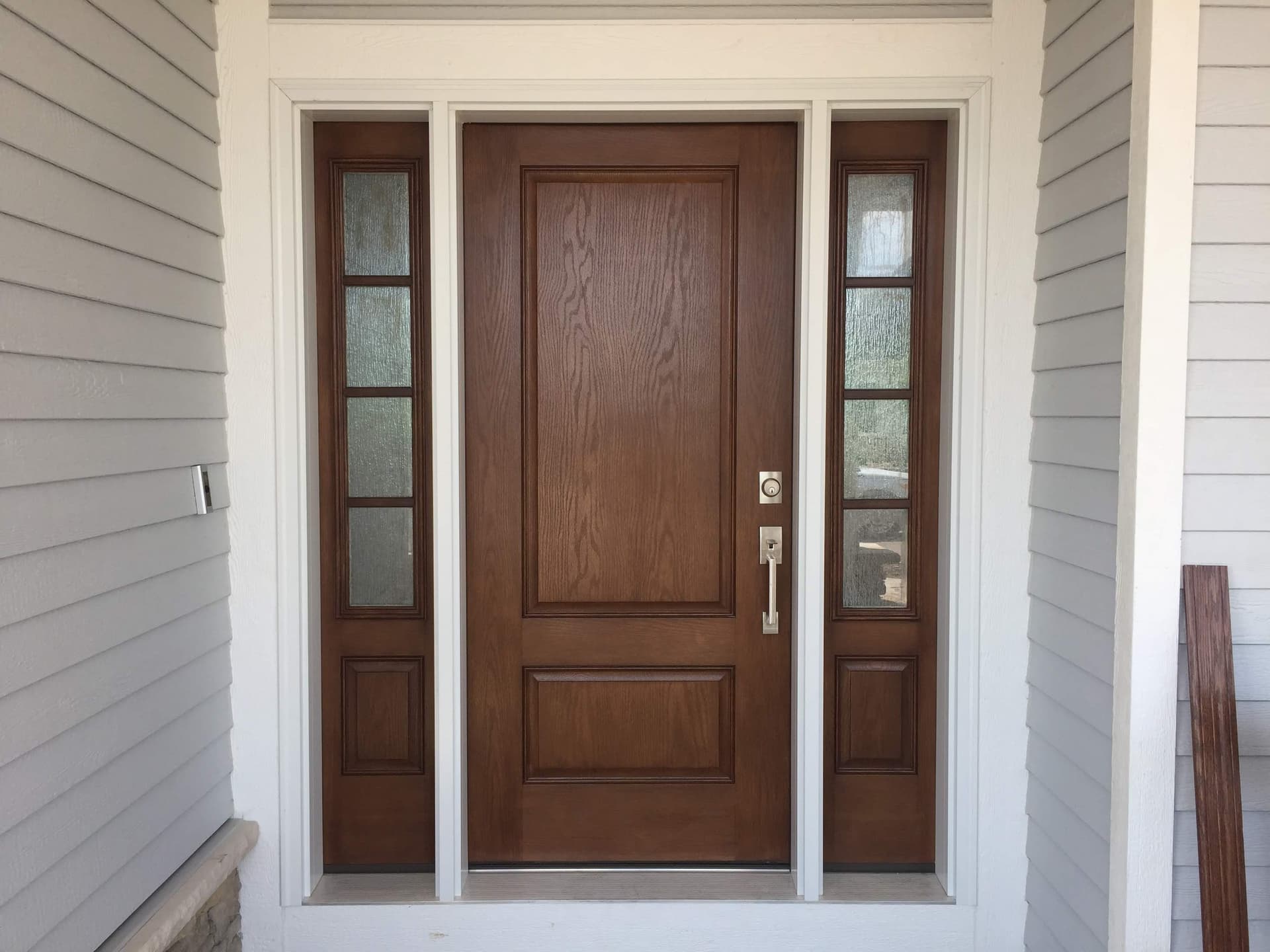 Brown-stained fiberglass entry door with two divided-light sidelights, centered in white lap siding with wide white casing.