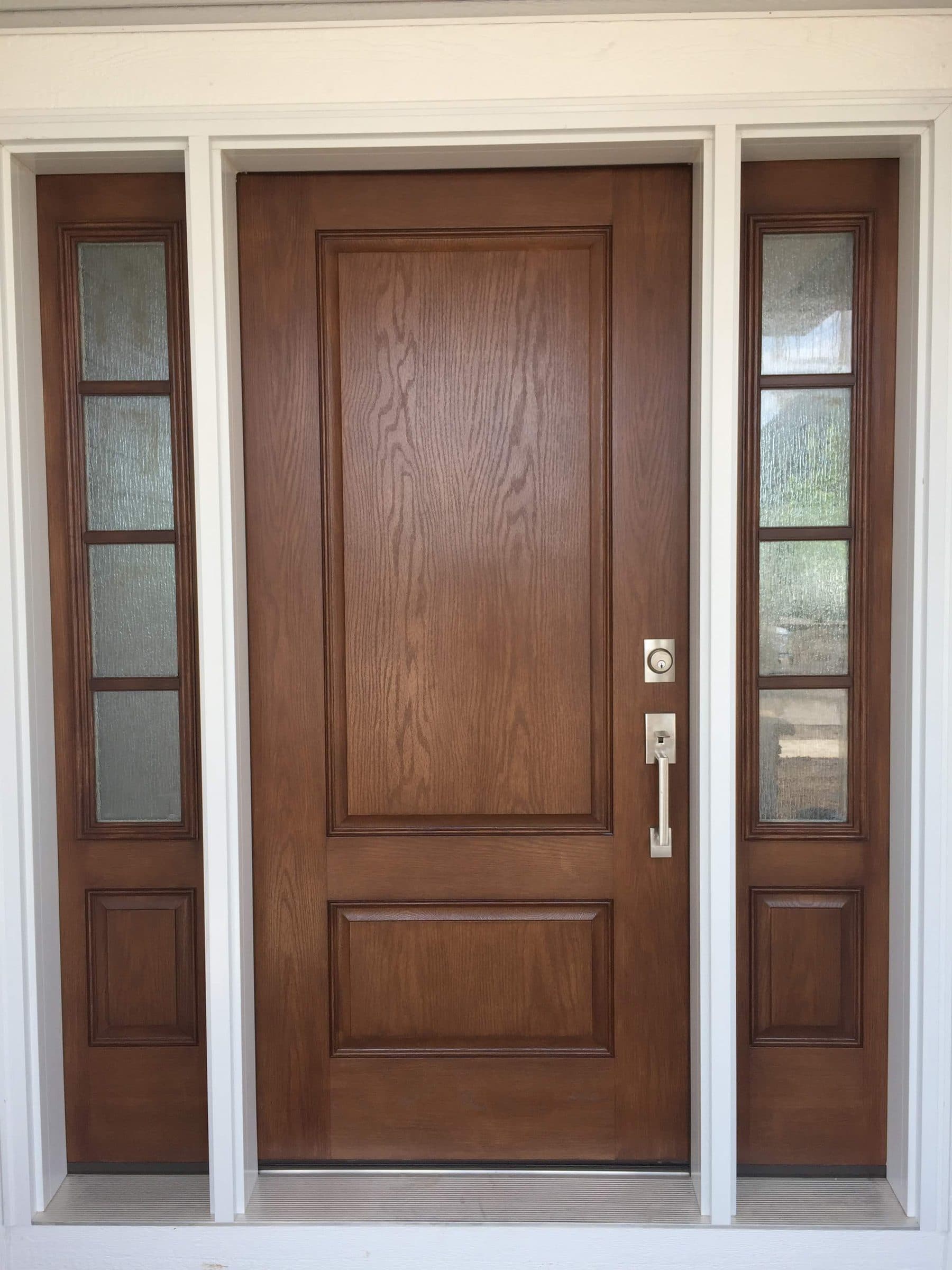 Brown-stained fiberglass entry door with pronounced oak graining and a brushed nickel handleset, flanked by two divided-light sidelights.