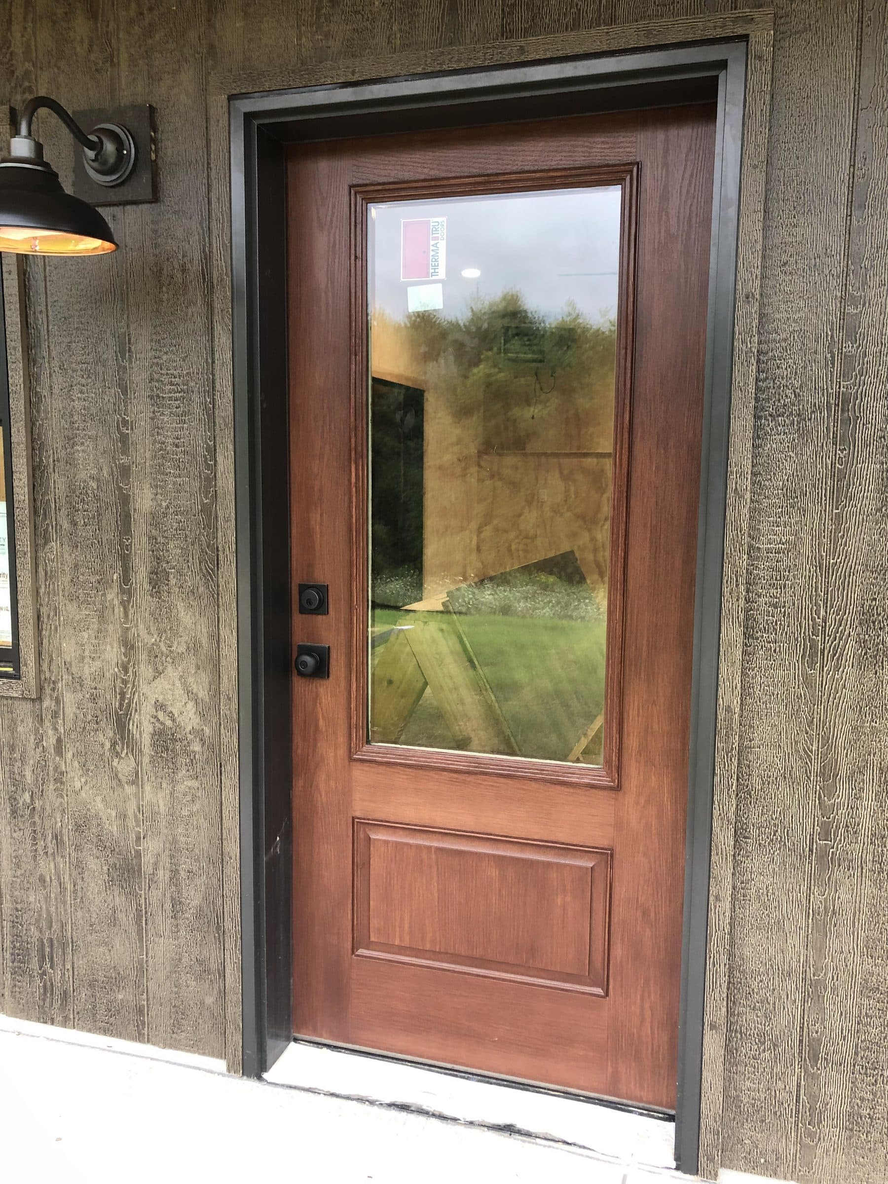 Brown-stained side entry door with a full-height glass panel under a black barn light, set in weathered gray board-and-batten siding.
