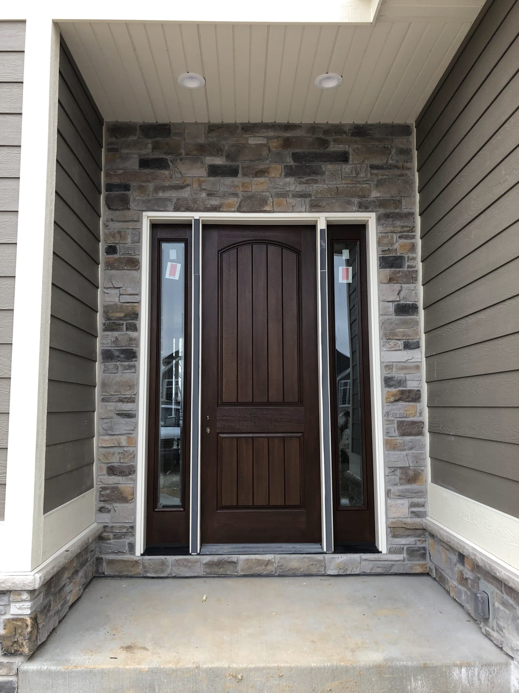 Wider view of the stained plank entry door from the covered porch, showing the stone veneer surround and the gray siding above.