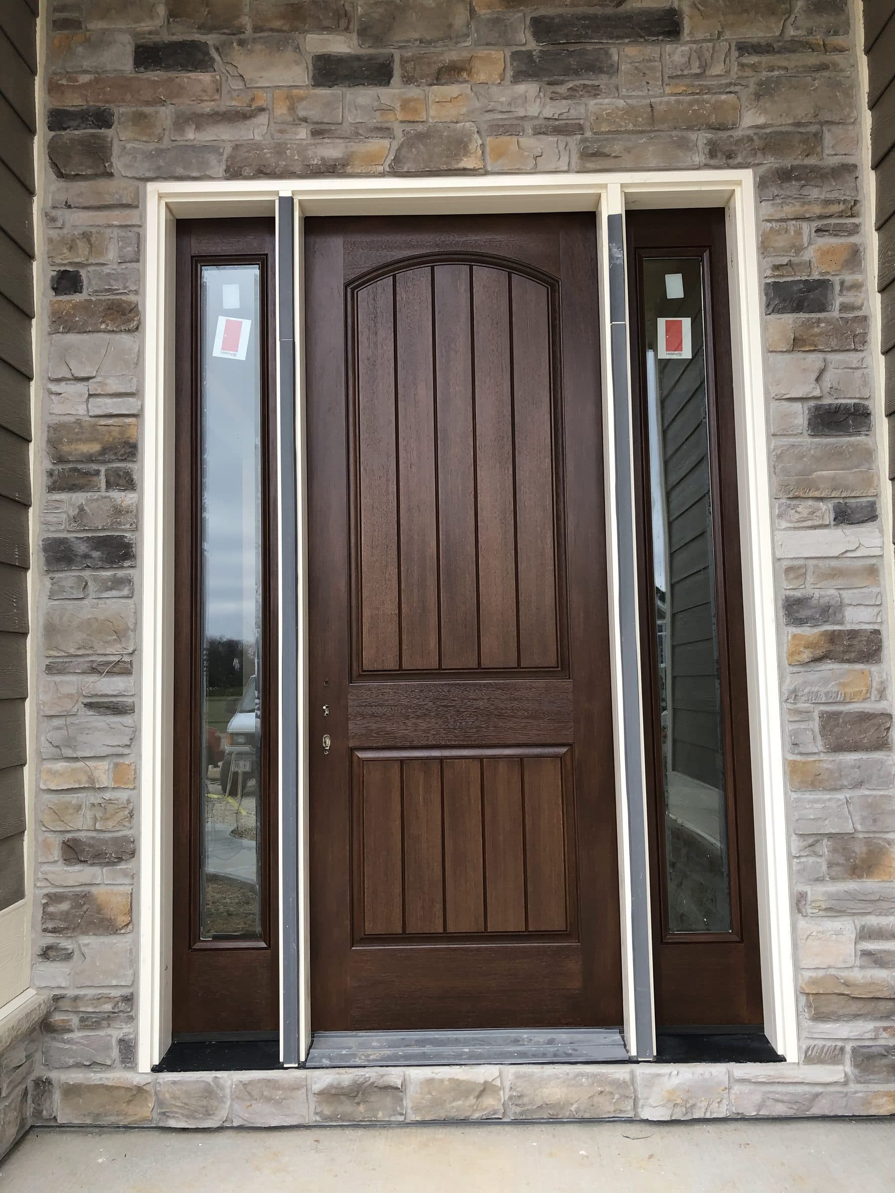 Brown-stained plank entry door with an arched top rail and glass sidelights, set into a mixed stone veneer wall.