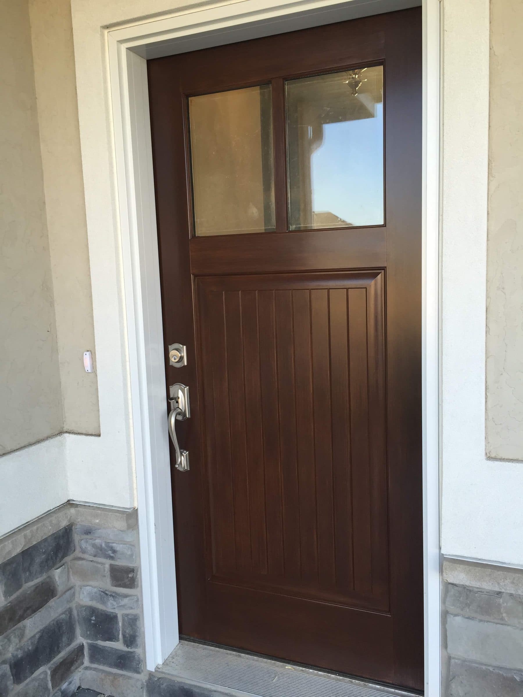 Brown-stained craftsman entry door with a single top light and a brushed nickel handleset, set in stucco above a gray stone base.