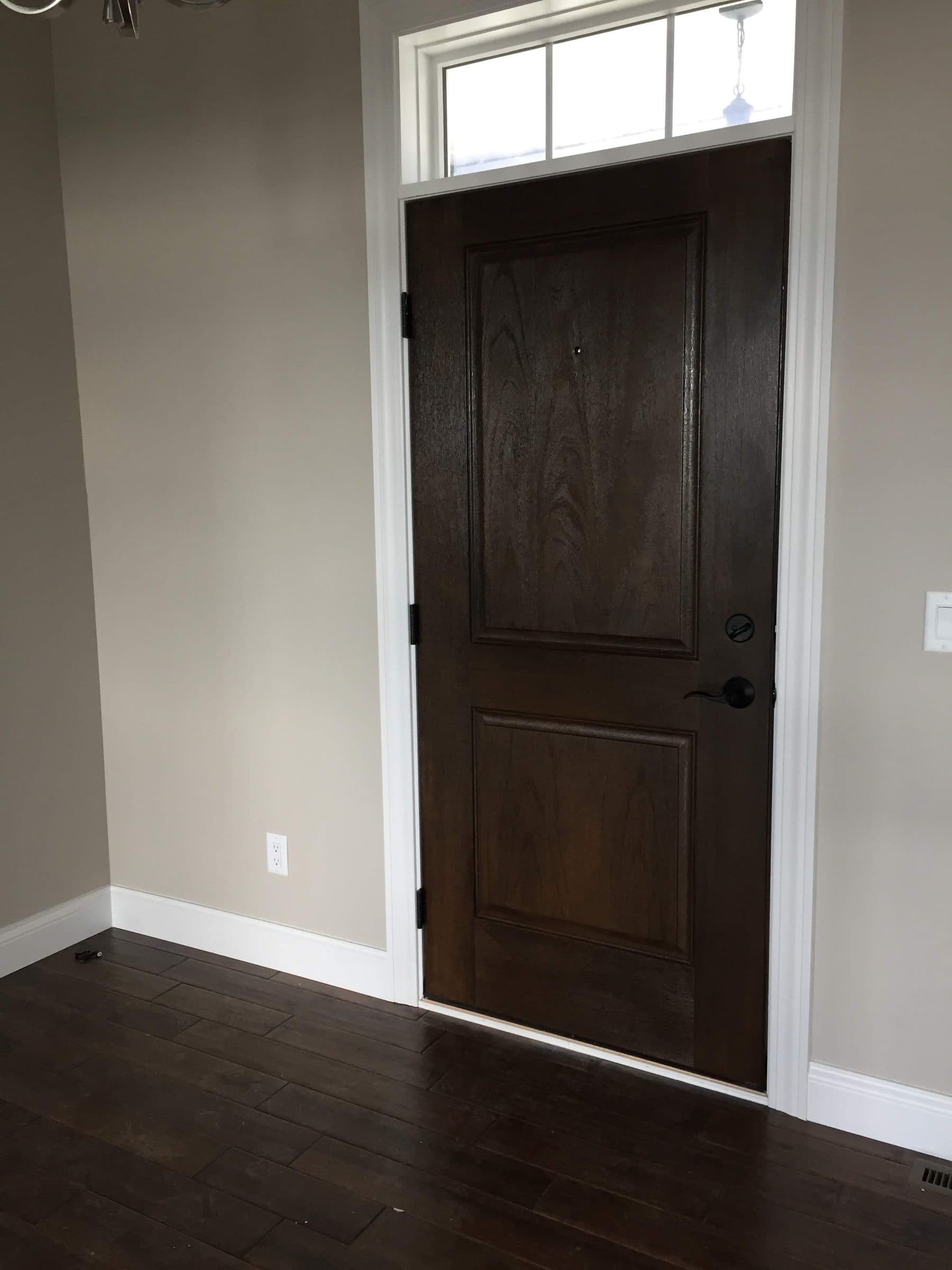 Dark walnut-stained entry door with a transom window above, seen from inside against white trim and dark hardwood floors.