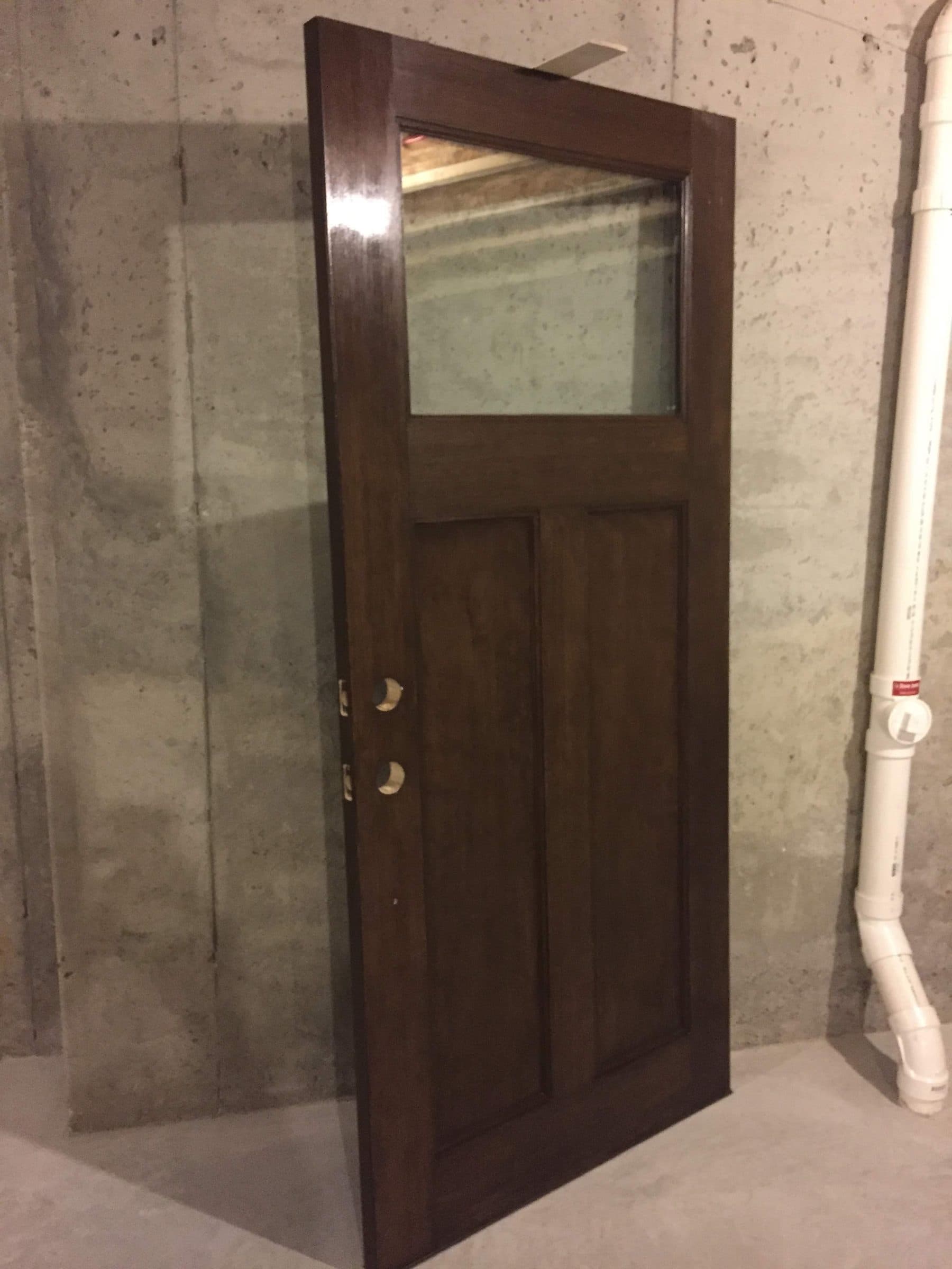 Refinished entry door standing off its hinges against a concrete foundation wall, showing the finished face and edges.