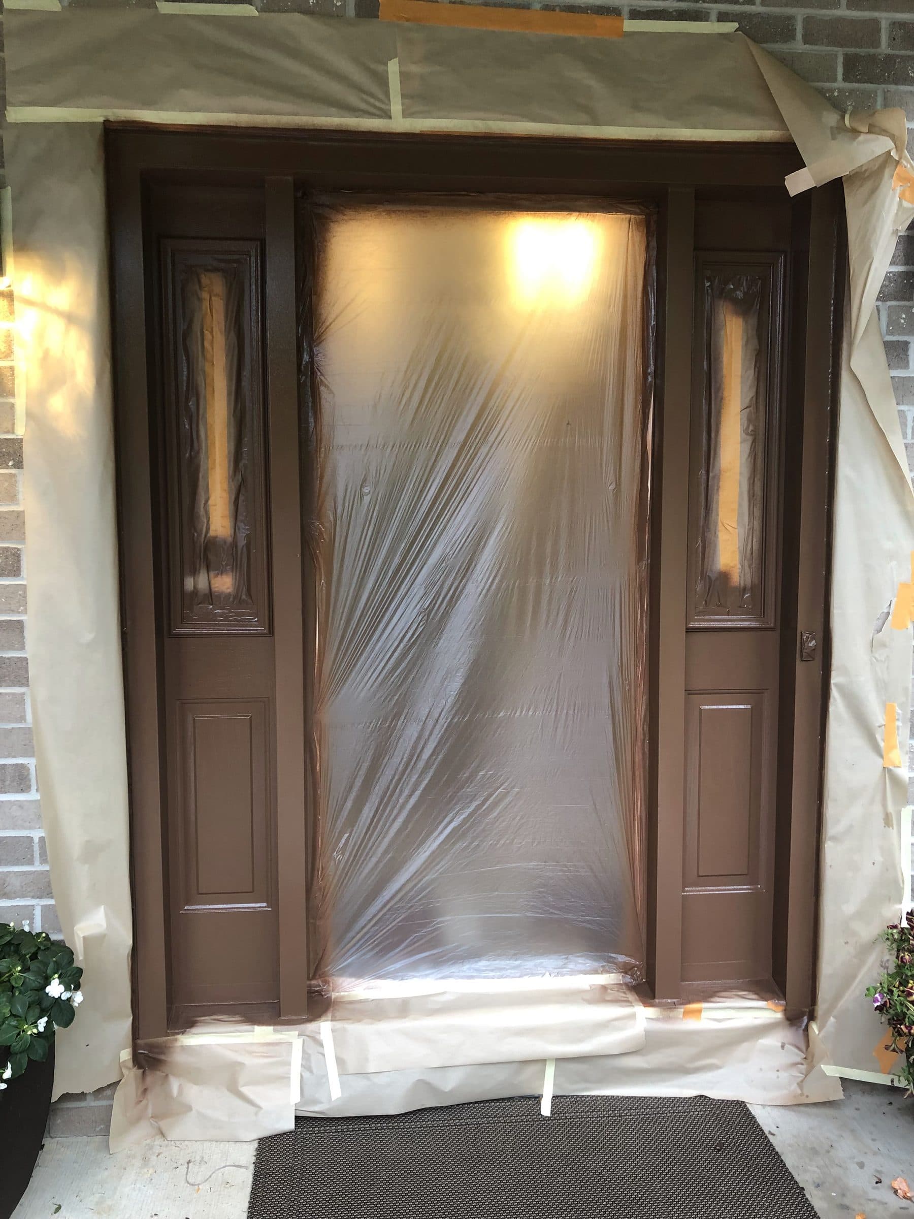 Brown entry door and sidelights fully masked in plastic sheeting and paper, with the floor covered, before staining begins.