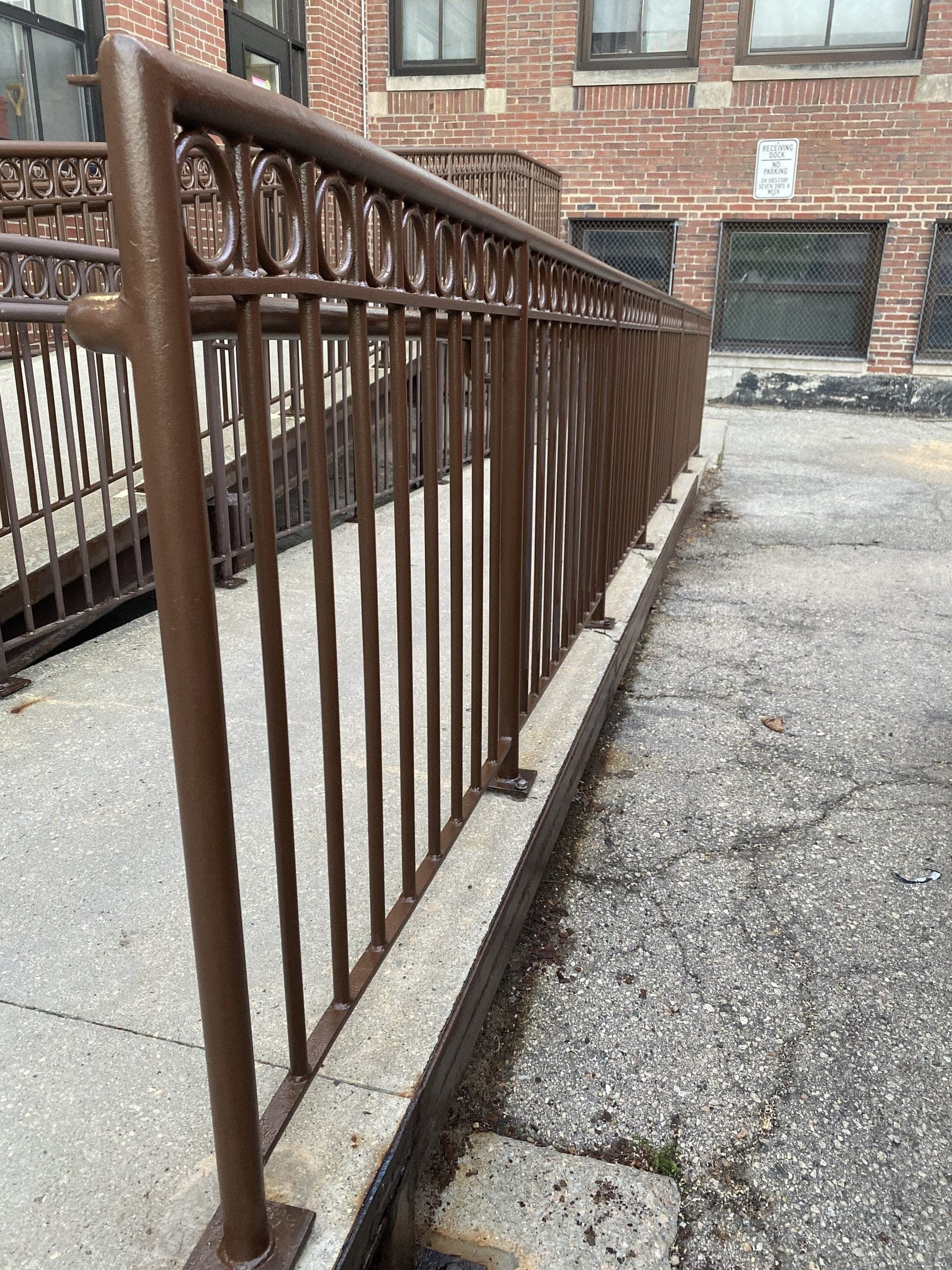 Bronze-brown iron handrail with a decorative ring detail running along a concrete walkway beside a brick building, de-rusted and recoated.