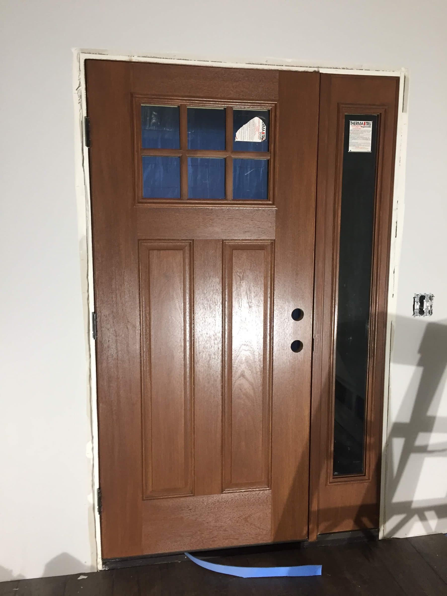 Fiberglass entry door part-way through staining, with blue tape masking the divided window panes and the sidelight beside it.