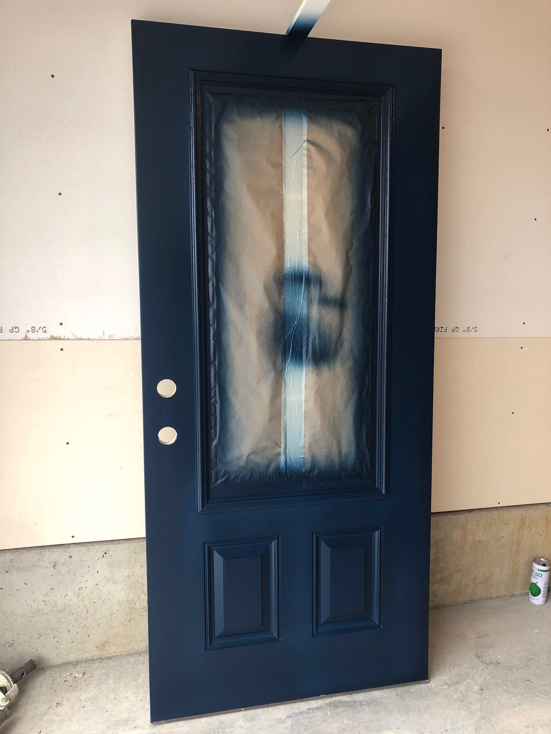Navy blue entry door taken off its hinges and stood up for spraying, with the glass masked and the panel edges coated.