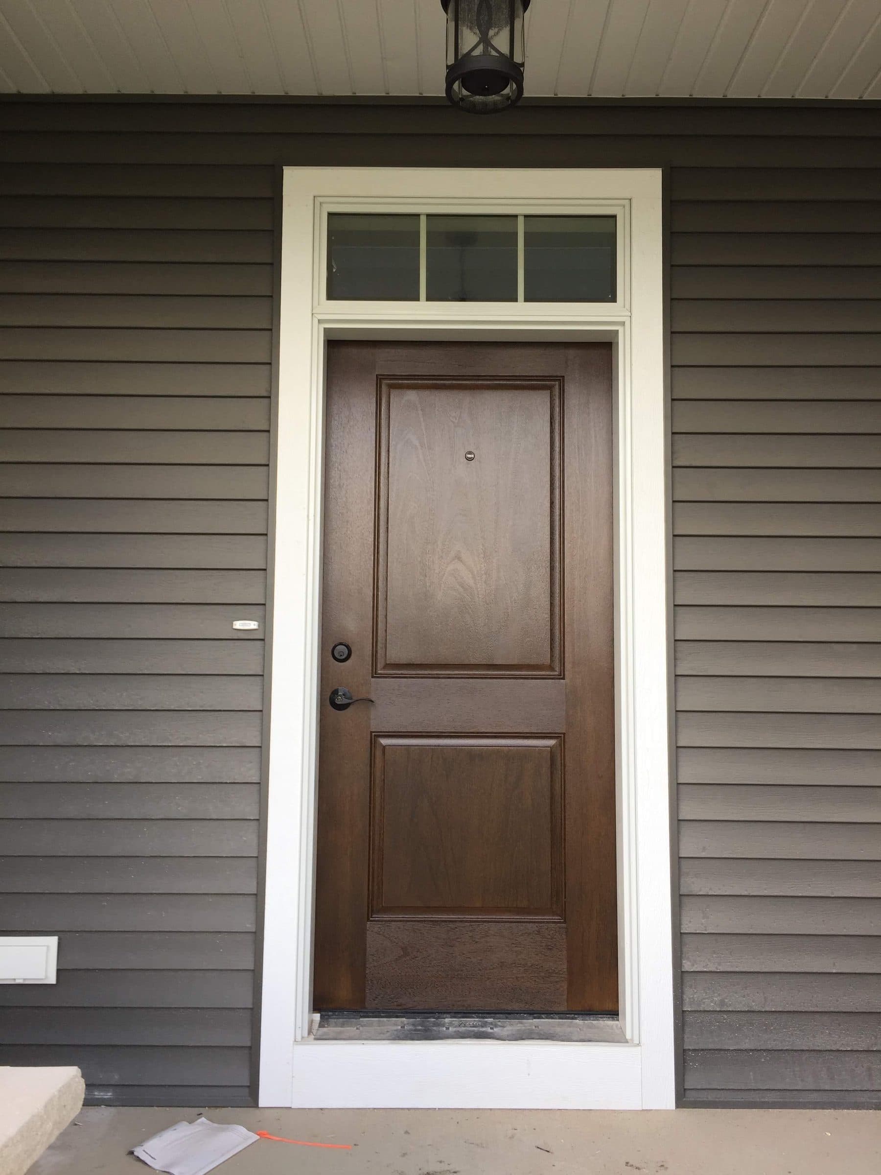 Brown-stained entry door with a divided transom above, set in gray lap siding with white casing.