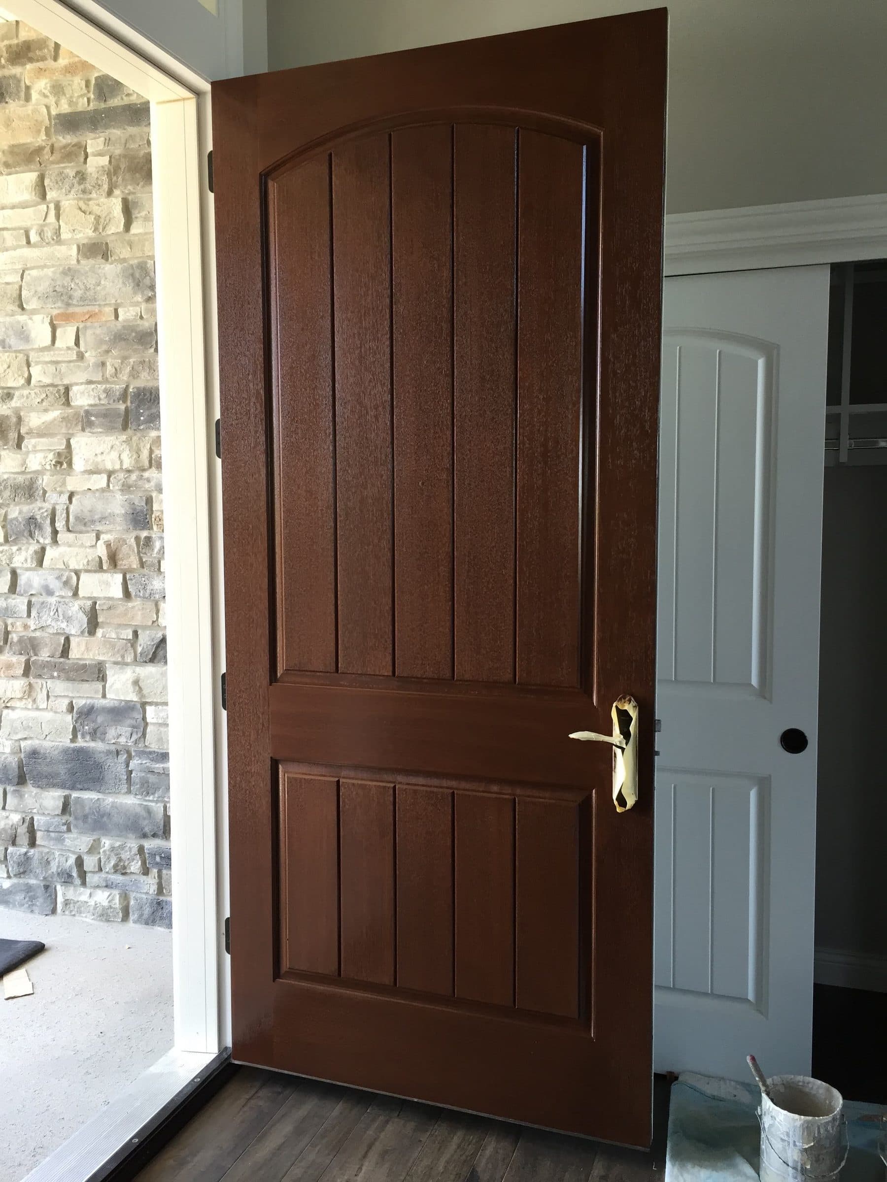 Brown-stained arched plank entry door standing open against a stone veneer wall, showing the finished hinge edge and the foyer beyond.