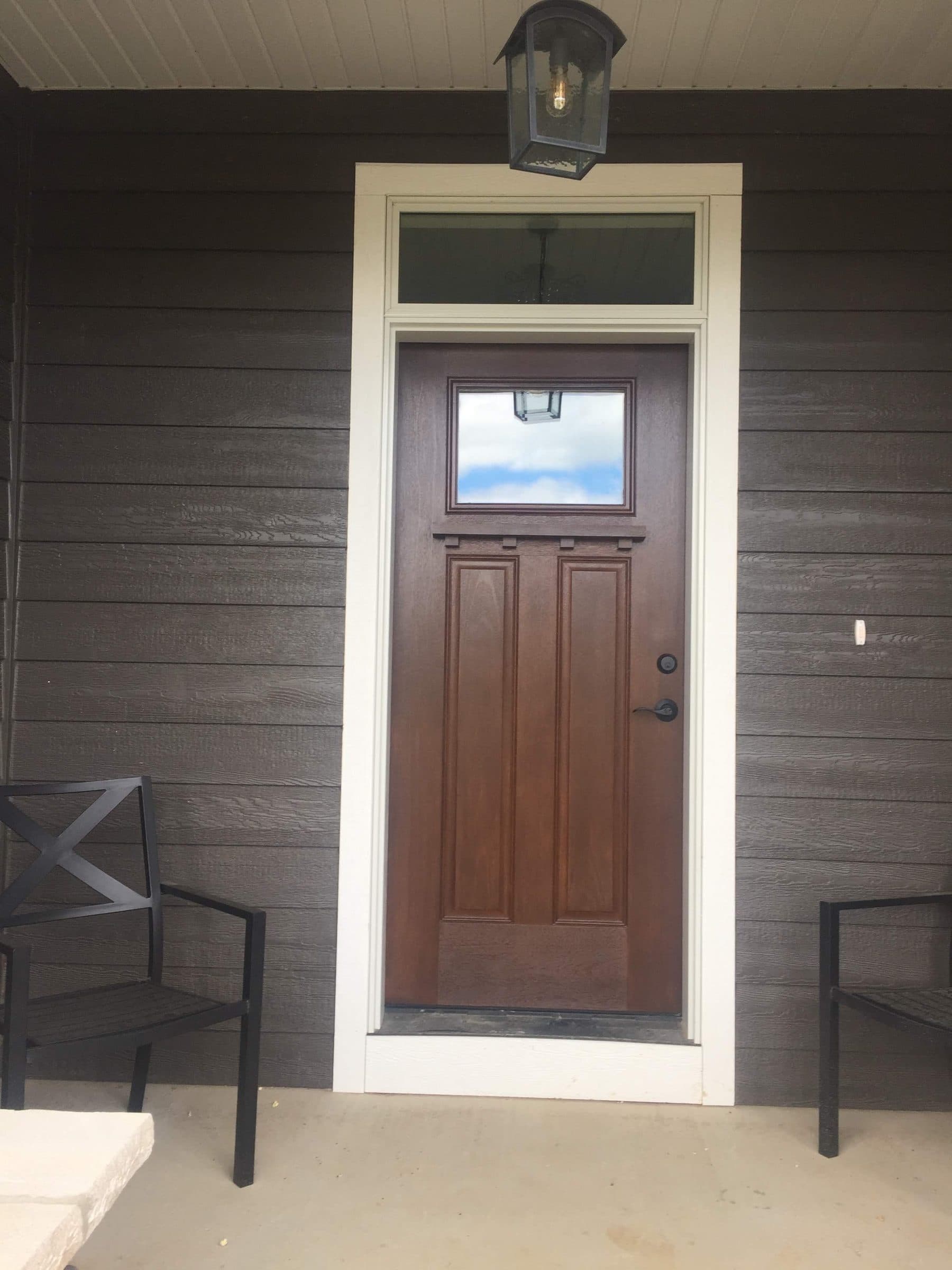 Brown-stained entry door with a single light and a transom above, set in dark brown siding beside patio chairs.