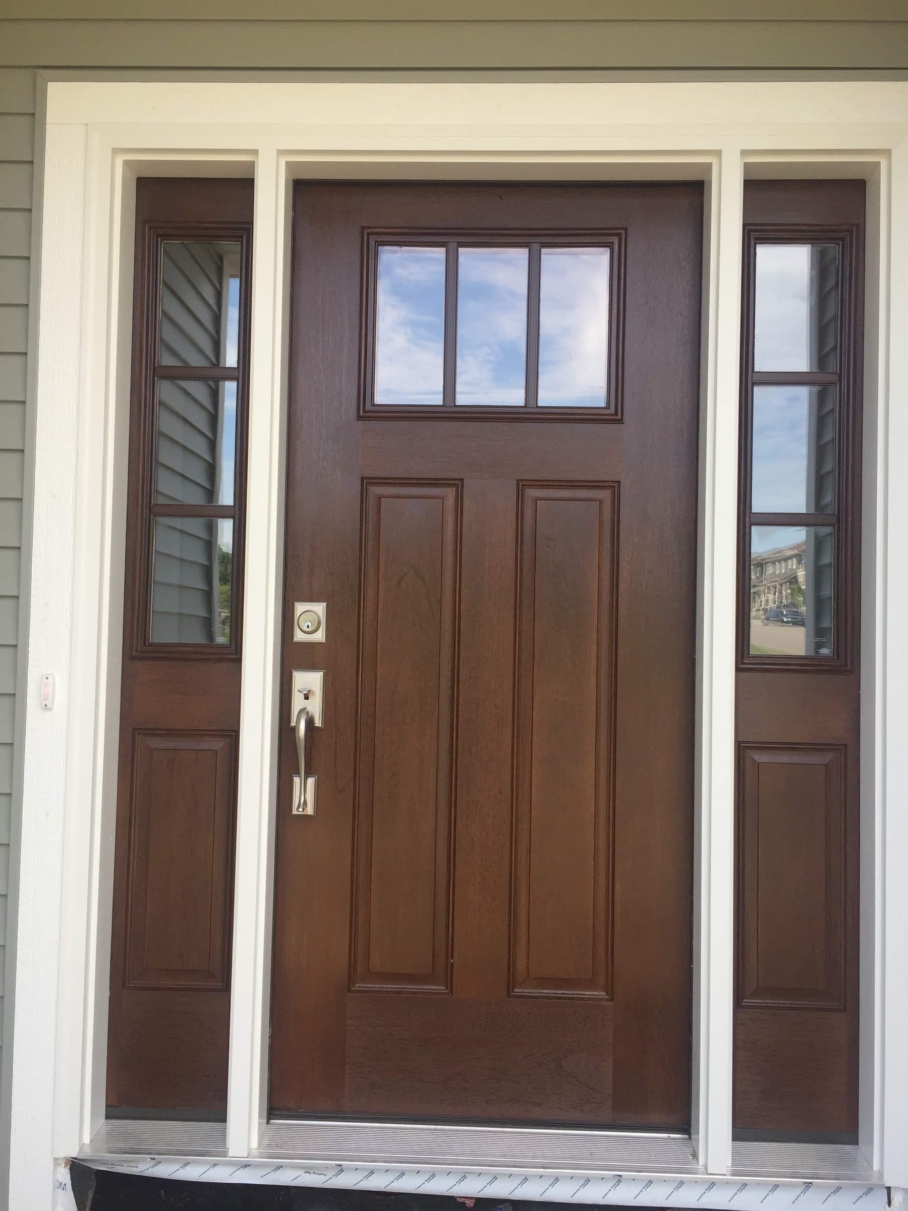 Brown-stained fiberglass entry door with a four-light top rail and two narrow sidelights, set in white lap siding.