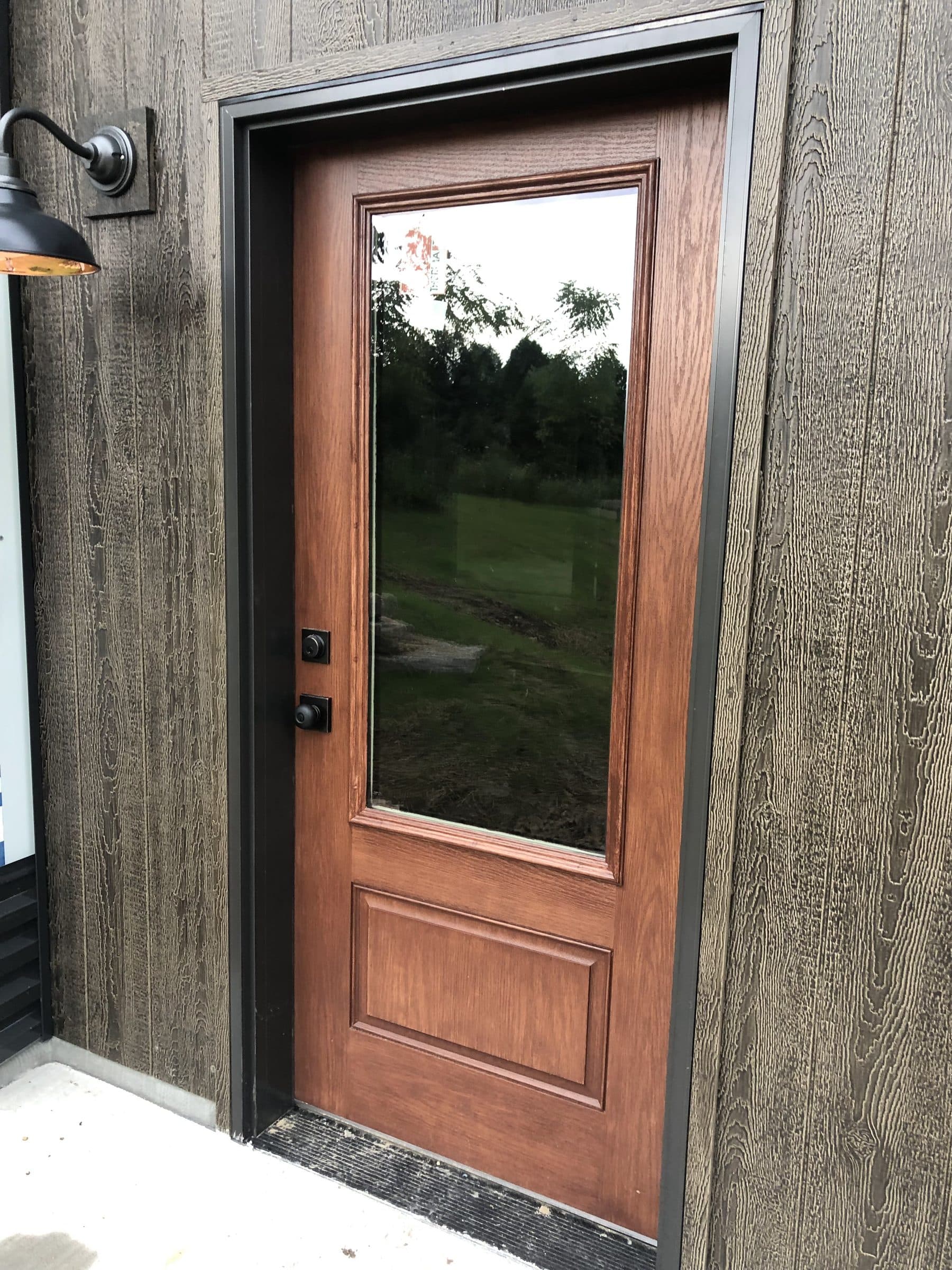 Brown-stained side door with a full-length glass panel under a black barn light, set in dark weathered siding.