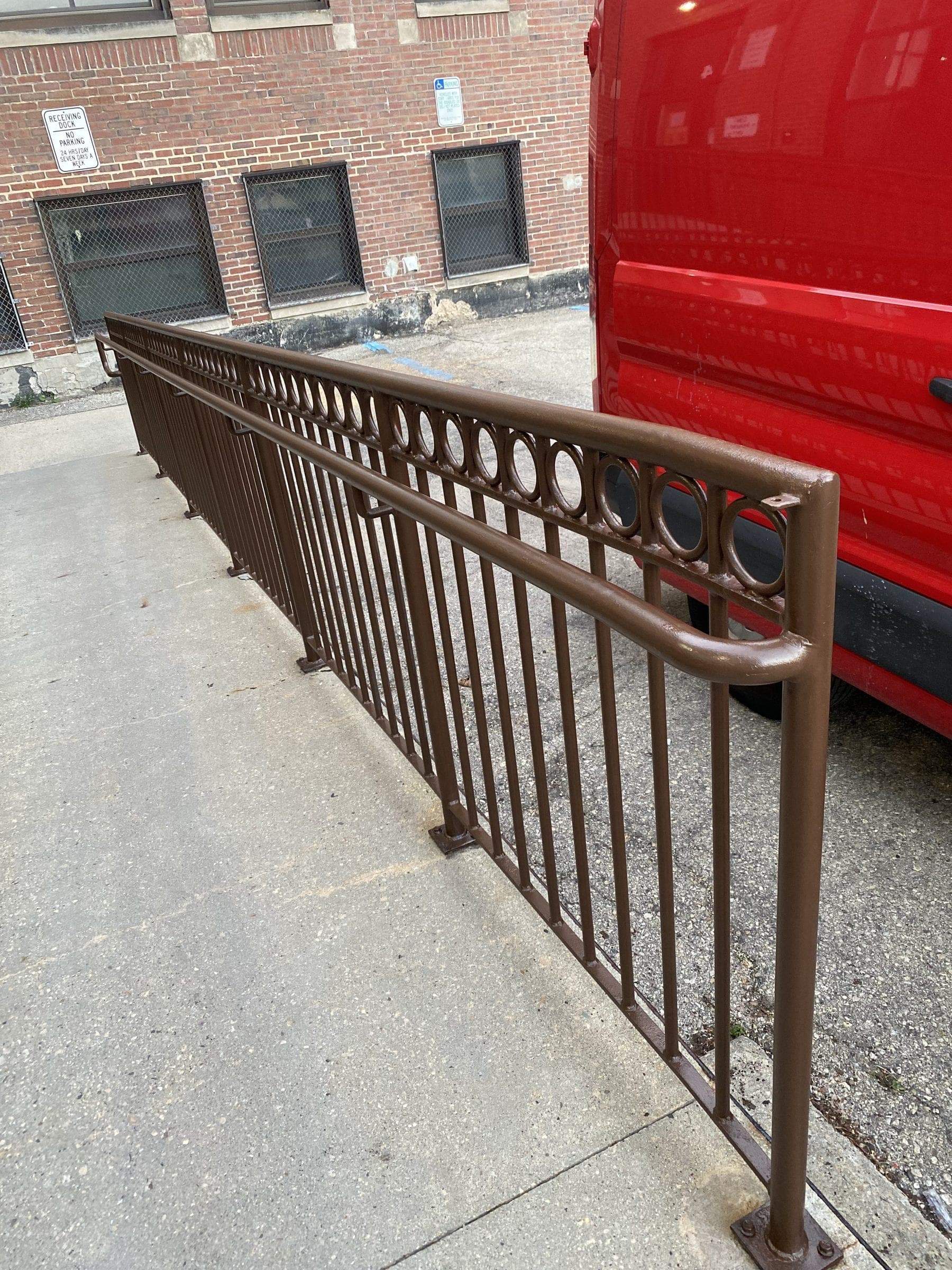 Long bronze-brown iron handrail with ring detail running the length of a concrete walkway outside a brick building.