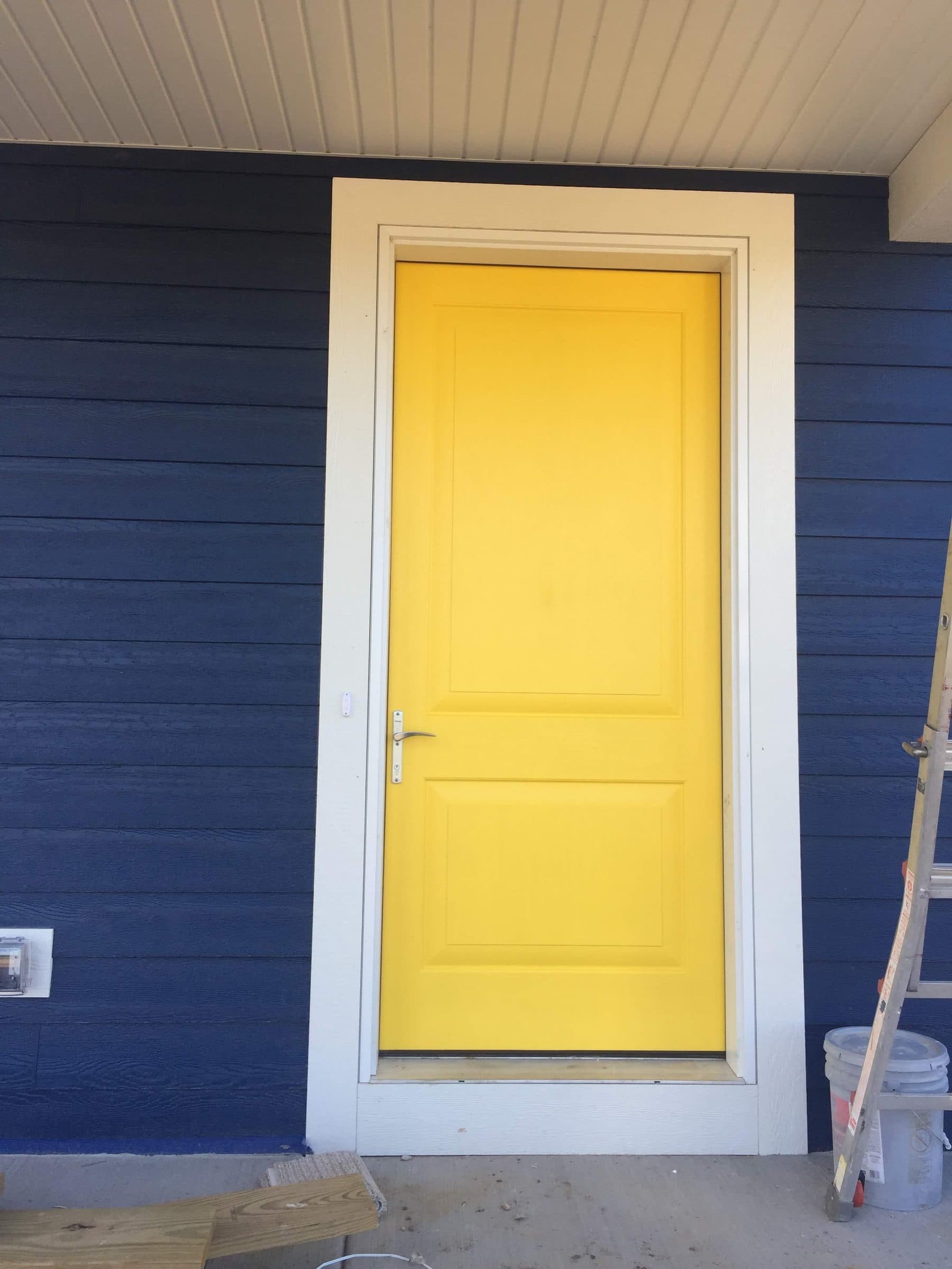 Bright yellow painted entry door in white casing against deep navy lap siding, with a transom light above.