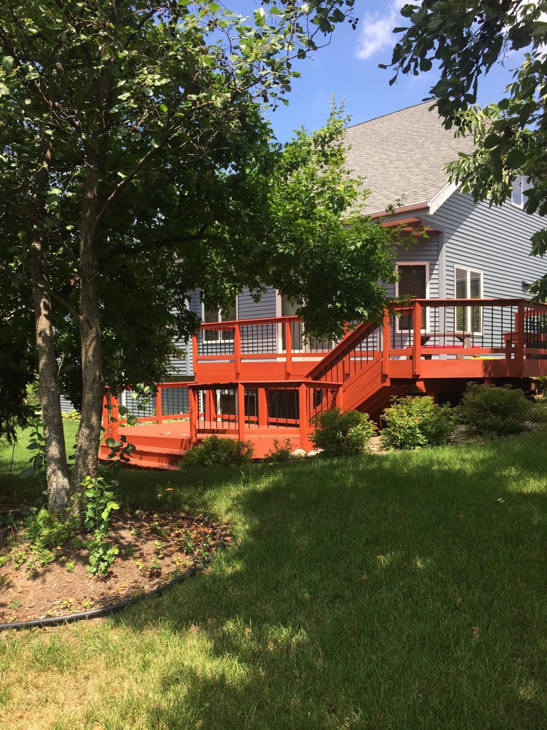 Two-level deck stained deep red against gray siding, seen across a mown lawn under mature trees.