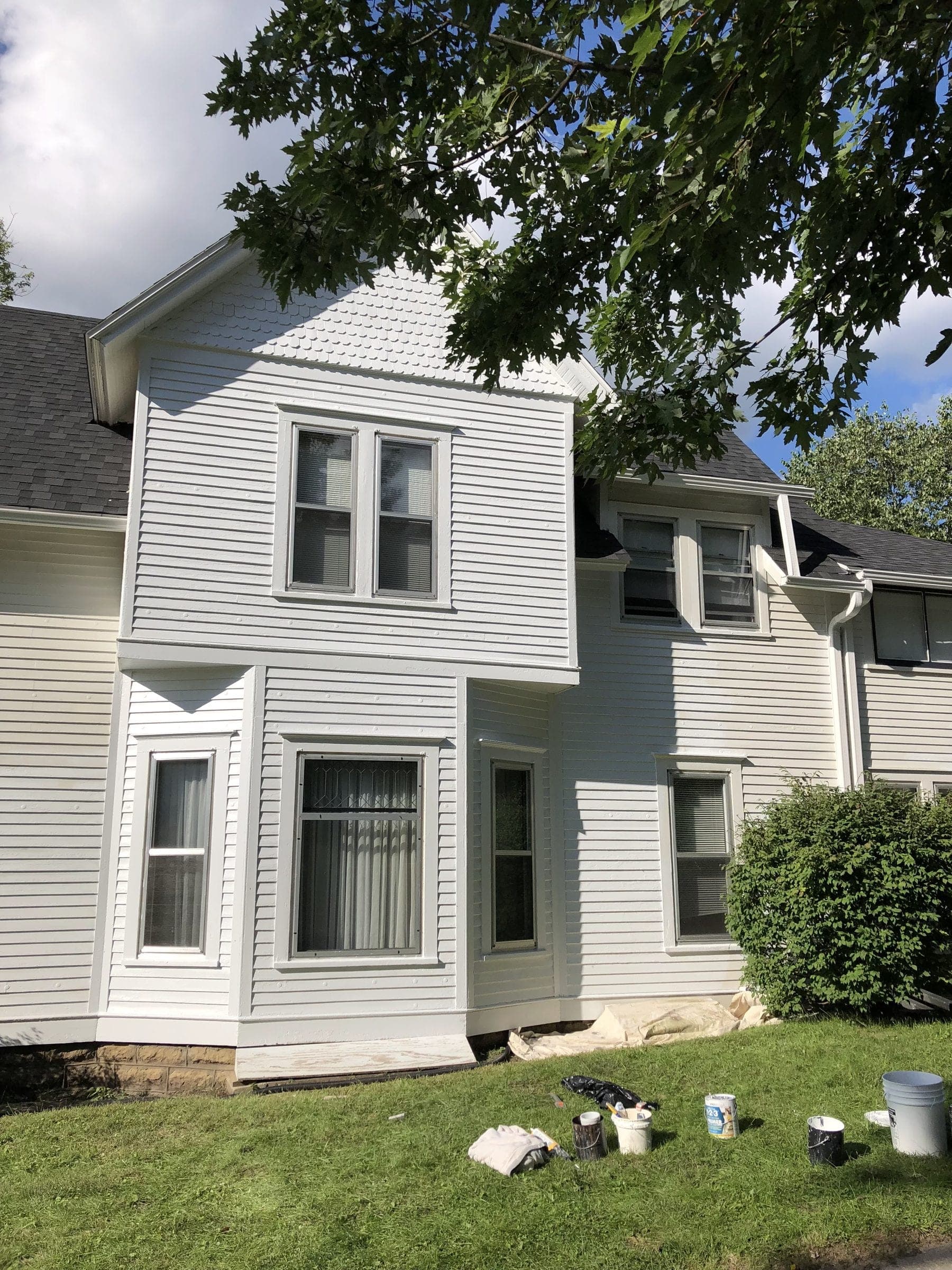 Two-story house repainted white with the bay window trim cut in, paint pails still standing on the grass.