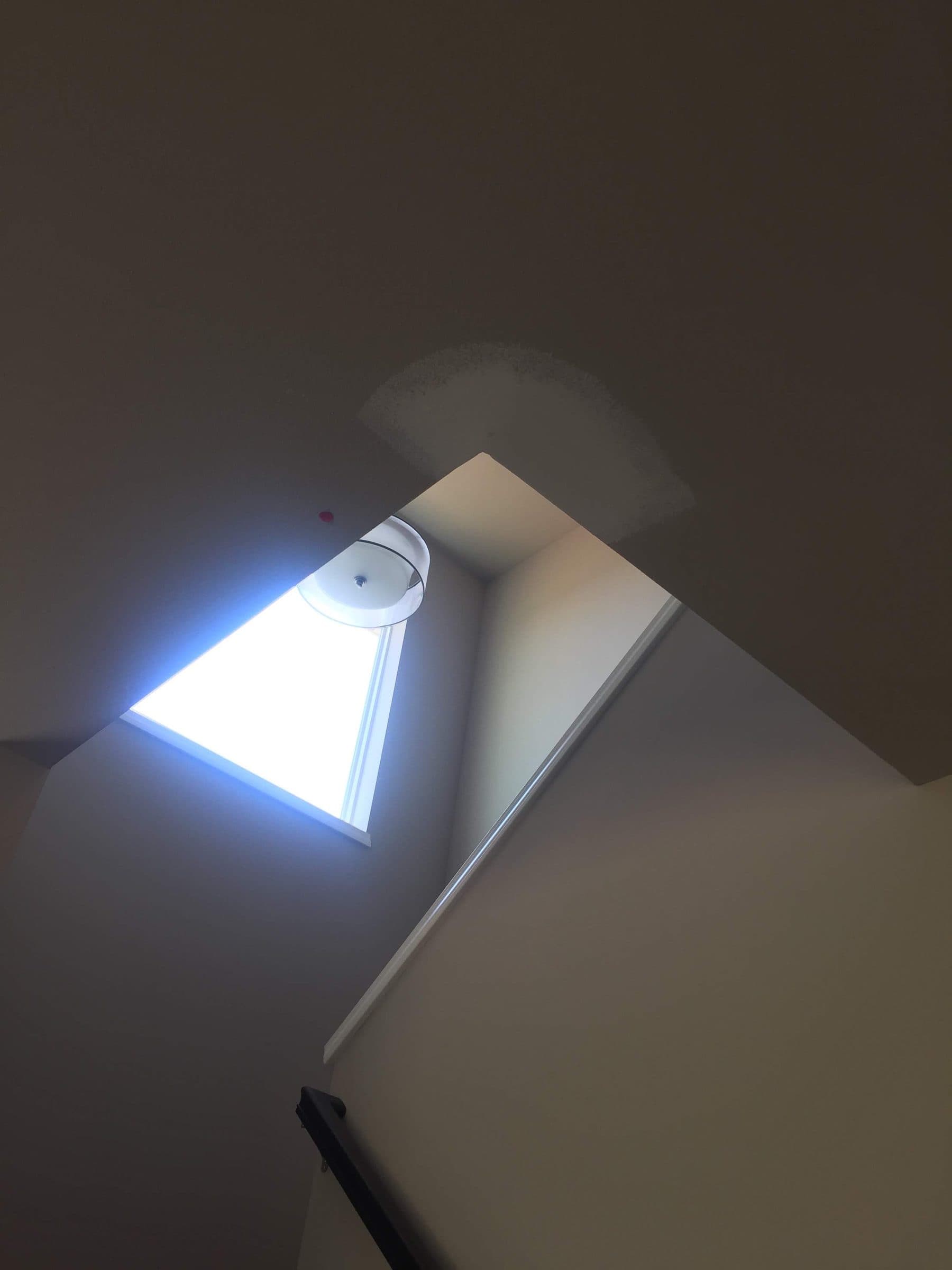 Drywall patch cut into a sloped ceiling beside a skylight, taped and skimmed ready for texture and paint.