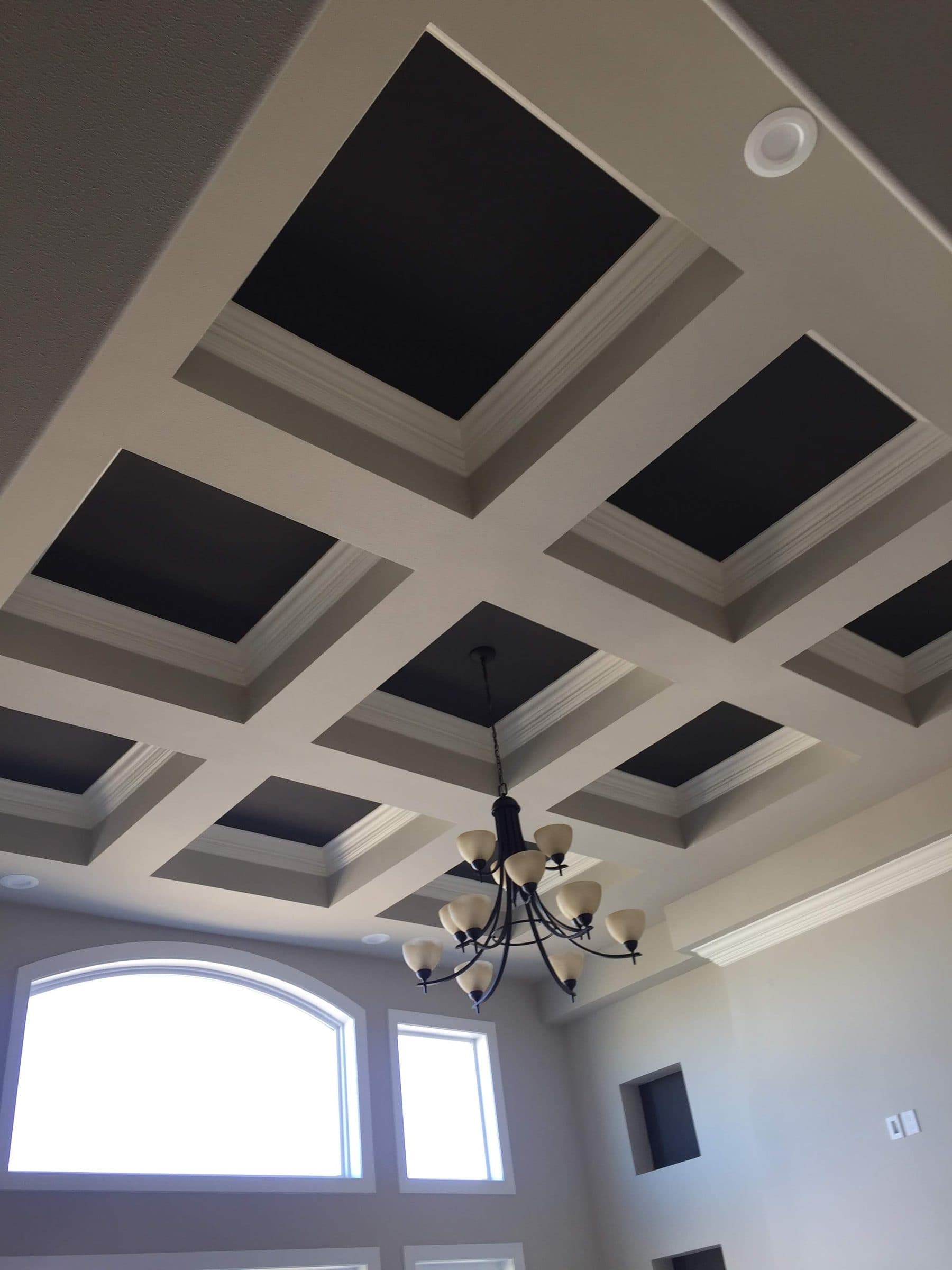 Coffered ceiling with black-painted recesses and pale gray beams, lit by an arched window wall and a black chandelier.