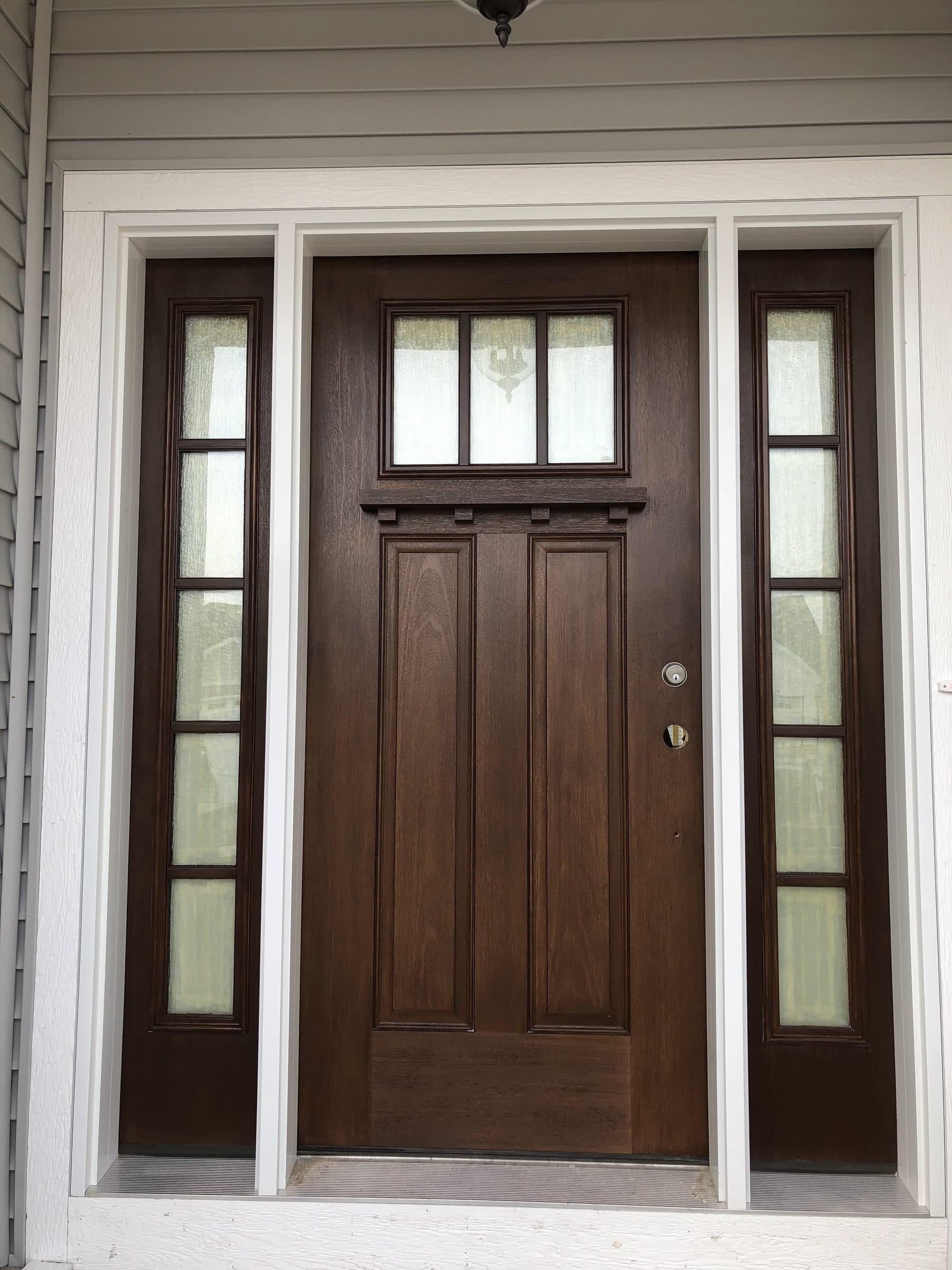 Craftsman-style entry door stained deep brown with a four-light window across the top rail, flanked by tall divided sidelights against gray siding.