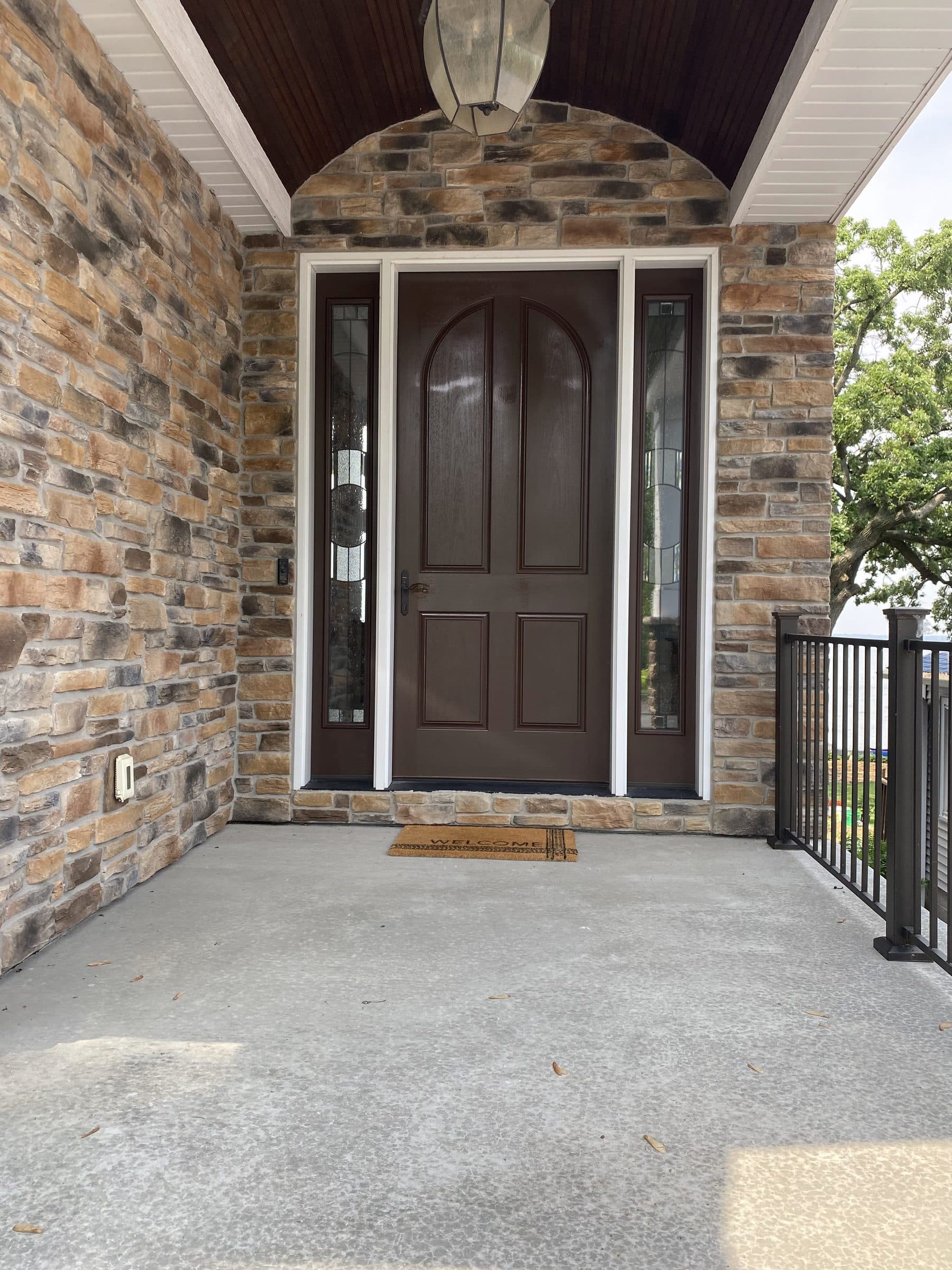 Dark-stained arched entry door with sidelights, finished and rehung under a bead-board porch ceiling with stone veneer walls.