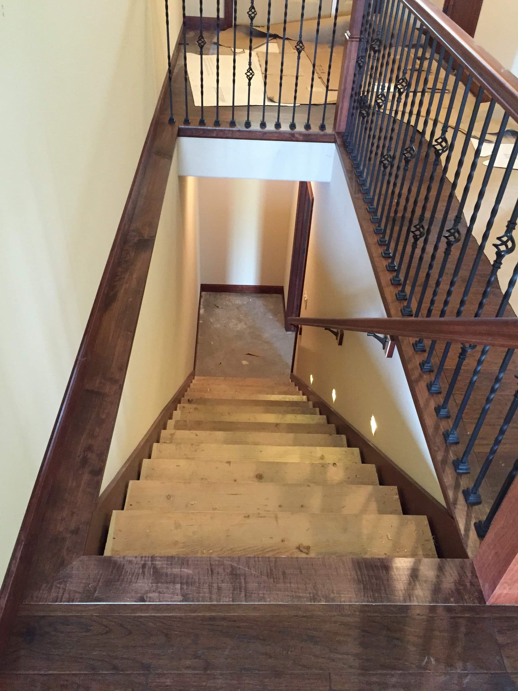 Stained handrail and newel with black iron balusters, seen looking down a staircase to the landing below.